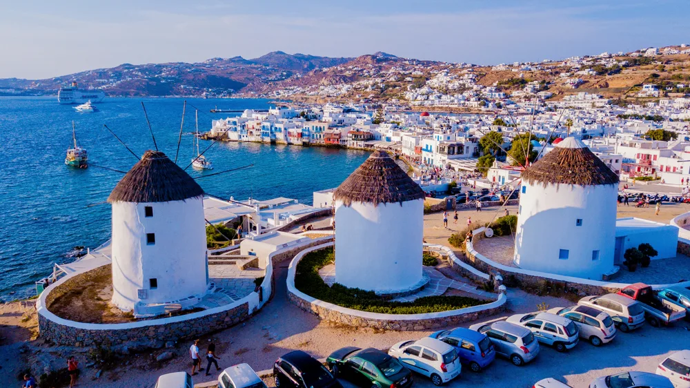 Three white native windmills near the coast with cars parked beside it, at a distance is coastal town, an image for a travel guide about safety in visiting Mykonos.