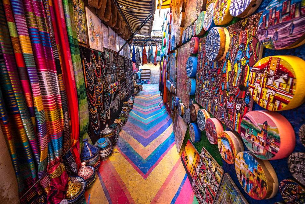 A small alley with a painted street and colorful artworks on round canvas are hanging on the walls, with vibrantly painted jars are placed on the opposite side.