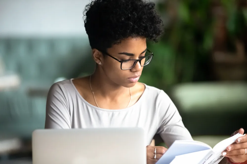 Young woman with a short curly hairdo concentrates at her laptop while flipping through a training booklet on how to become a travel agent