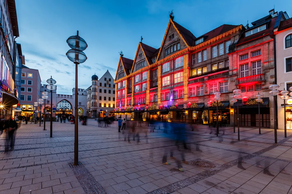 People can be seen walking in a town square where a structure is decorated by lights during dusk, a long exposure image for a travel guide about safety in visiting Munich.