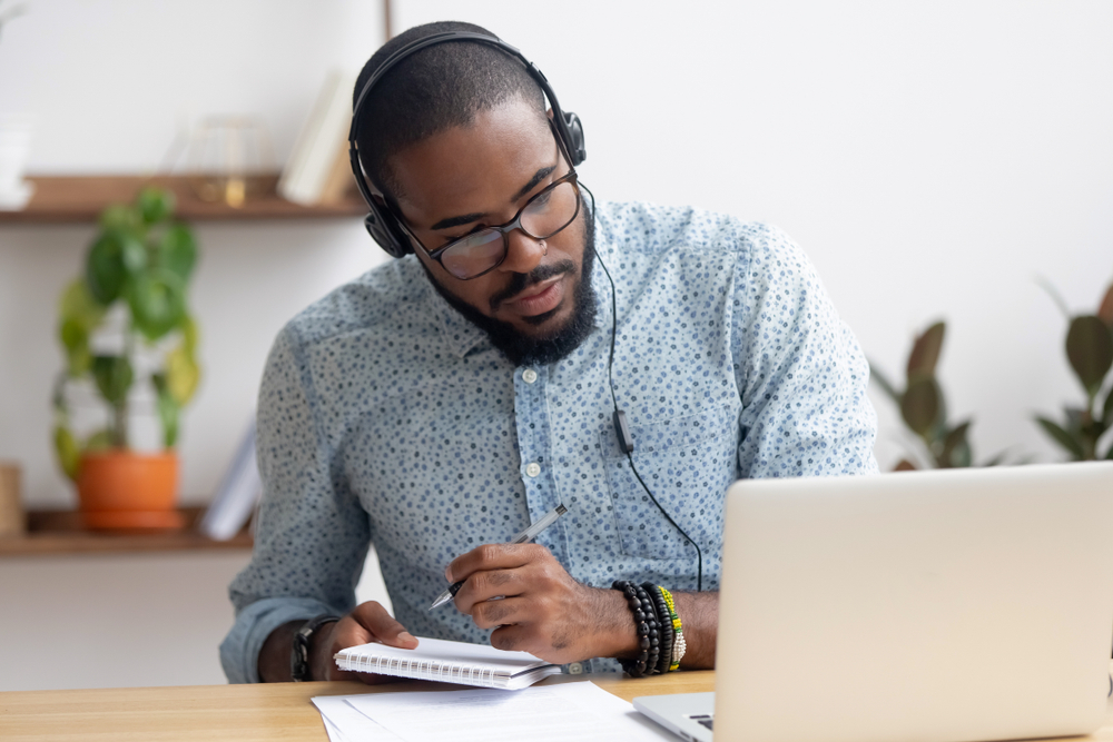 Young African American man studies to become a travel agent for free while taking notes in front of his laptop with headphones on
