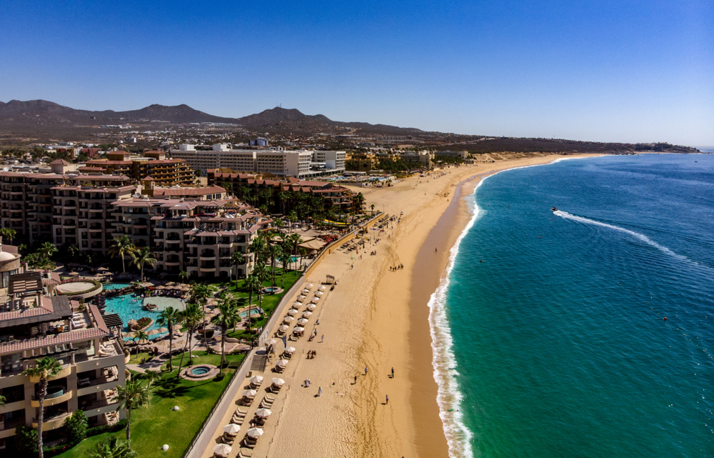 Aerial image of a bunch of hotels lining the sandy shore of Medano Beach pictured for a guide titled Where to Stay in Cabo