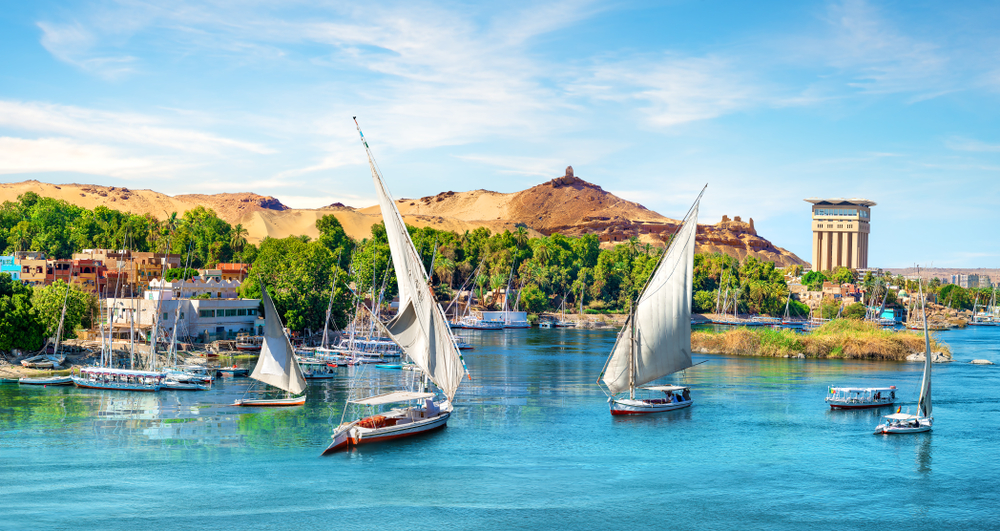 Sailboats floating on large river at Aswan, pictured or a piece on where to stay in Egypt, other boats are docked on a pier with lush trees.