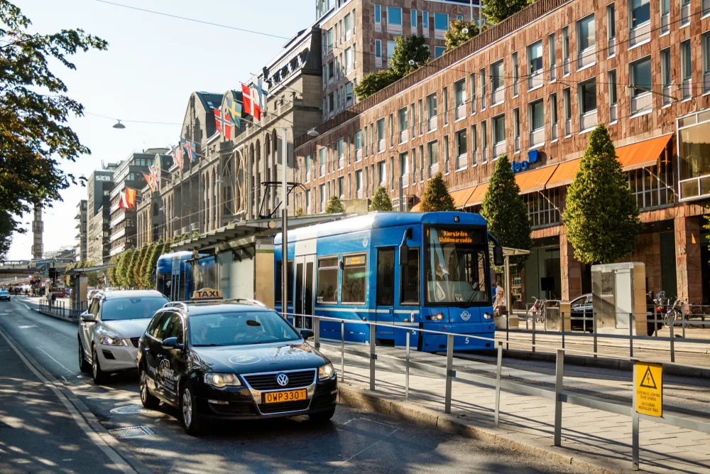 Featured as an image for a guide titled Is Sweden Safe to Visit, a photo of a blue train and taxis with modern and historic buildings in the background with trees lining the tracks and a bridge in the background
