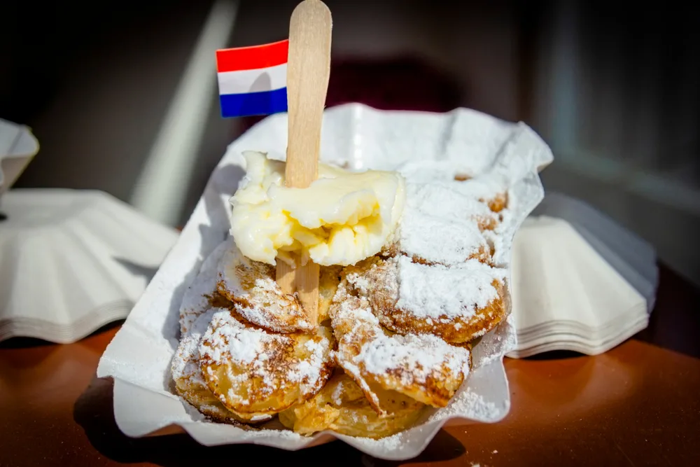 Fresh poffertjes mini pancakes dusted with powdered sugar and topped with butter and a Dutch flag from a street vendor shown as one of the best Dutch foods to try