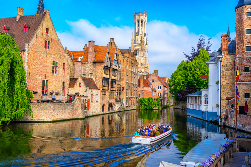 A group of tourist riding a small boat  cruising on a canal in between old structures in the city, an image for a travel guide about trip cost to Belgium. 
