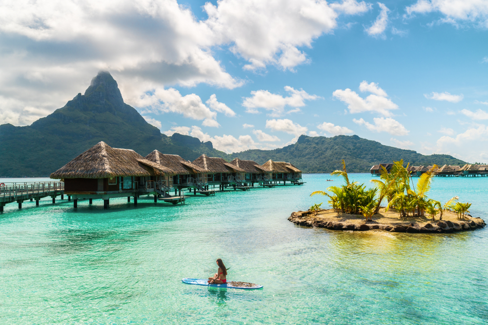 A woman paddle boarding on crystal clear waters beside a series of native houses interconnected by a boardwalk in the middle of the beach, and a tall mountain is in background, a section image for an article about trip to Tahiti cost.