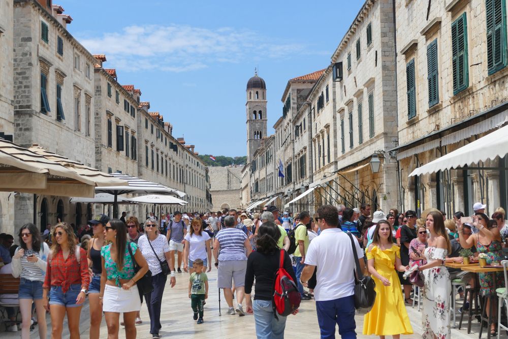 Photo of a crowded city street for a guide titled Is Dubrovnik Safe to Visit with a blue sky above the walled town and its many cafes