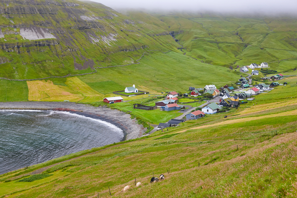 A small coastal village sits at the valley, where grass-covered fields can be seen. 