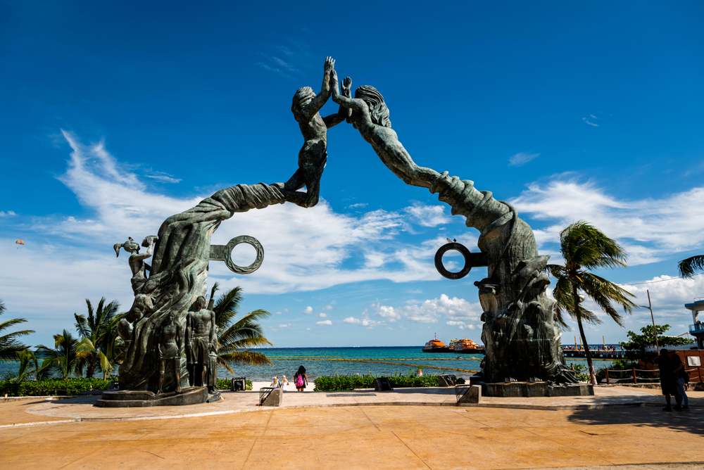 A huge statue of two people pushing against each other while standing on a vine-like structure, and other smaller statues stand at the bottom of it. 