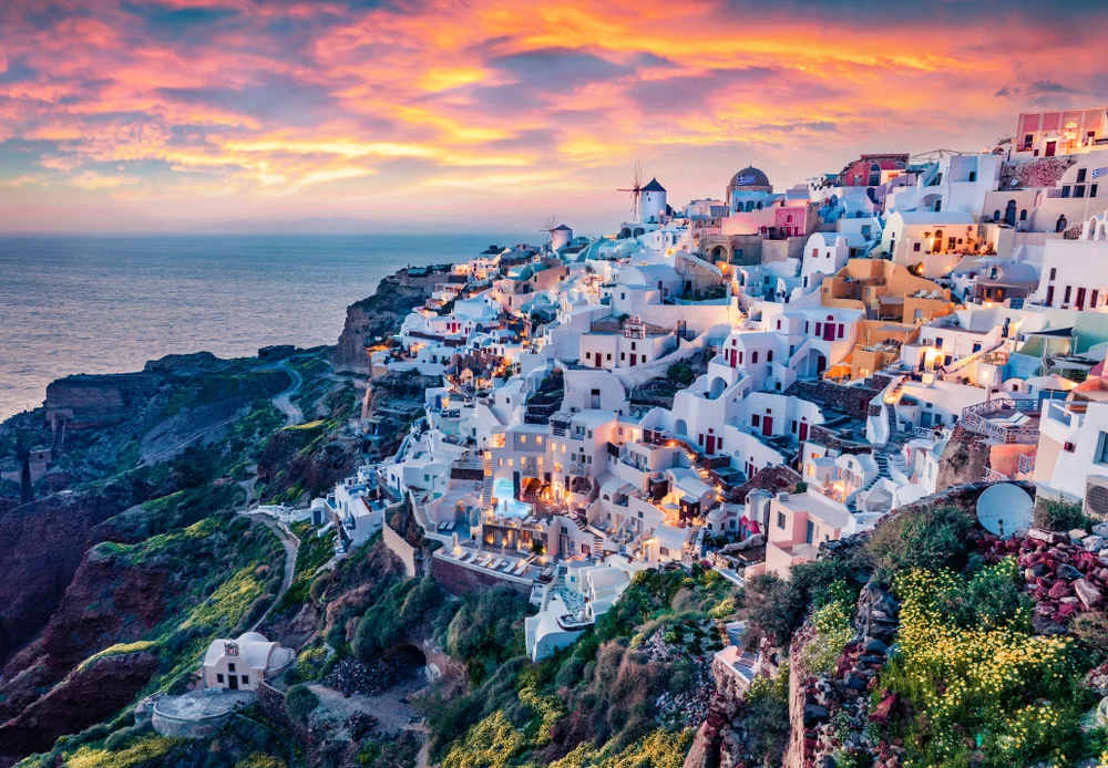 Oia, Santorini in Greece shown at sunset with buildings on the hill lit up to show what the Greek party islands look like at sunset