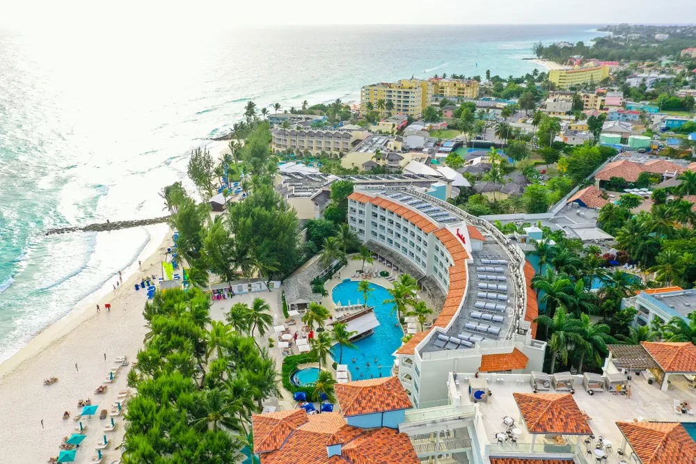 Aerial view on a resort with a swimming pool beside the beach in Oistins, one of the best areas to stay in Barbados, and a few tourists and sun beds on the white shore. 
