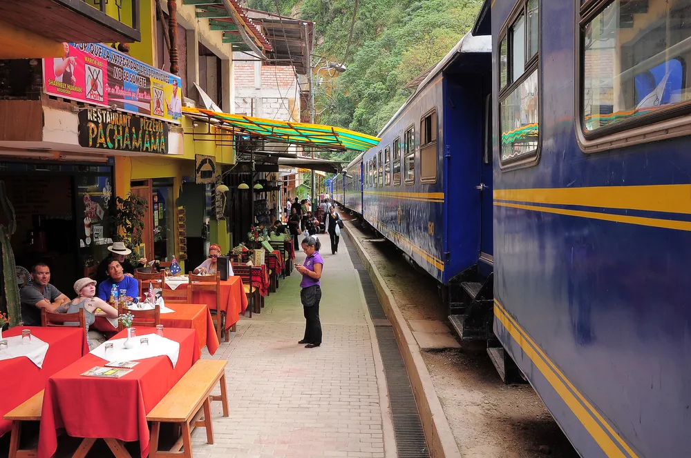 People dining at restaurants during a train stop, an image for a travel guide about trip cost to Machu Picchu, a blue train is standby on its tracks.