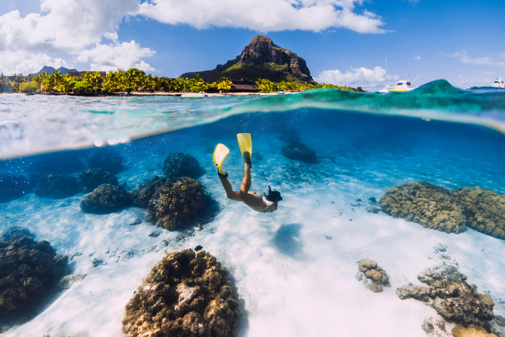A woman diving wearing a swimming wear and fins in a crystal clear sea, and on the top parts of the photo, the land area can be seen with a mountain.