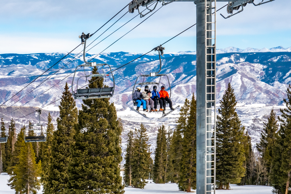 Ski lift with people riding it up the mountain at Aspen's Snowmass Ski Resort during winter with mountains in the distance