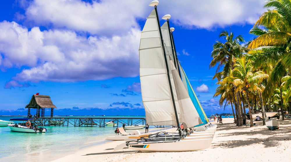 Sailboats on the white sand shore of Trou-aux-Biches and Pointe aux Canonniers, two of the best areas to stay in Mauritius, short boardwalk with a native structure extends from the shore with palm trees to the beach.