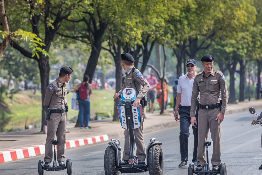 Three asian police mounted on their mobile vehicle as they are roving the street, and a tourist is seen walking behind them. 