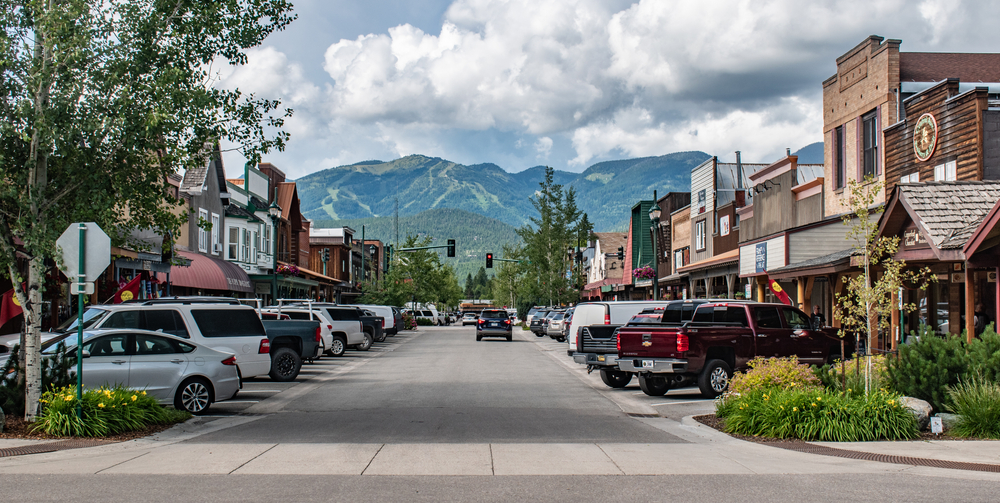 View of a town with cars parked in front of the structures and in background are tall mountains. 