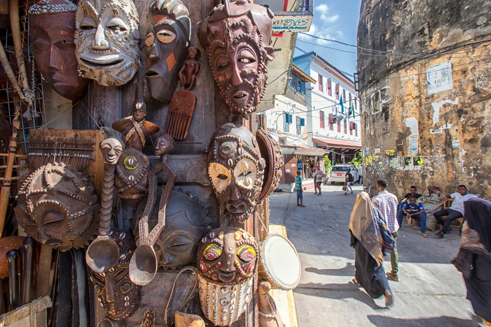Indigenous masks made of wood displayed on the streets of Stone Town, one of the best places to stay in Zanzibar, and people can be seen walking on the street of the town.