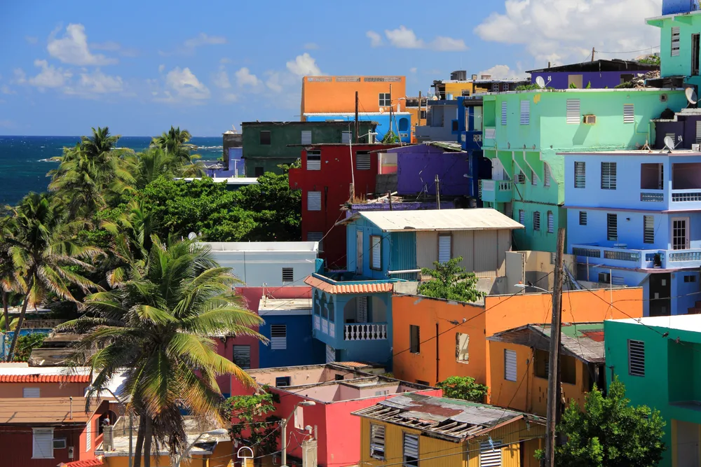 Photo of homes in the La Perla area pictured for a guide titled Is San Juan Safe to Visit featuring lots of colorful buildings on a hillside with a blue sky and ocean in the background