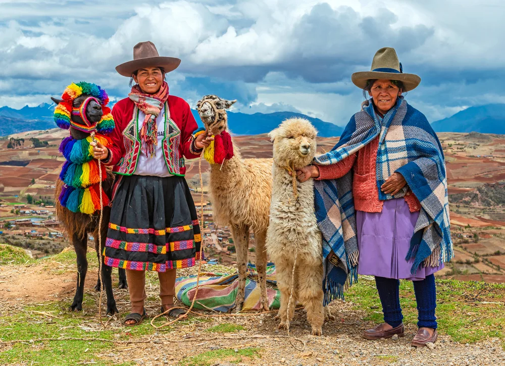 Two elderly local women wearing traditional clothing with their Alpacas on to high-altitude location, an image for a travel guide about trip cost to Peru. 