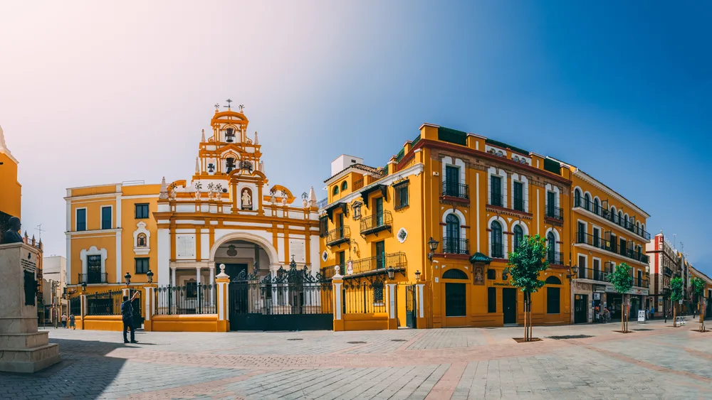 A panoramic view of a series of old buildings in La Macarena, a historic experience on the best areas to stay in Seville, a person stands in front of the white church beside a yellow building.
