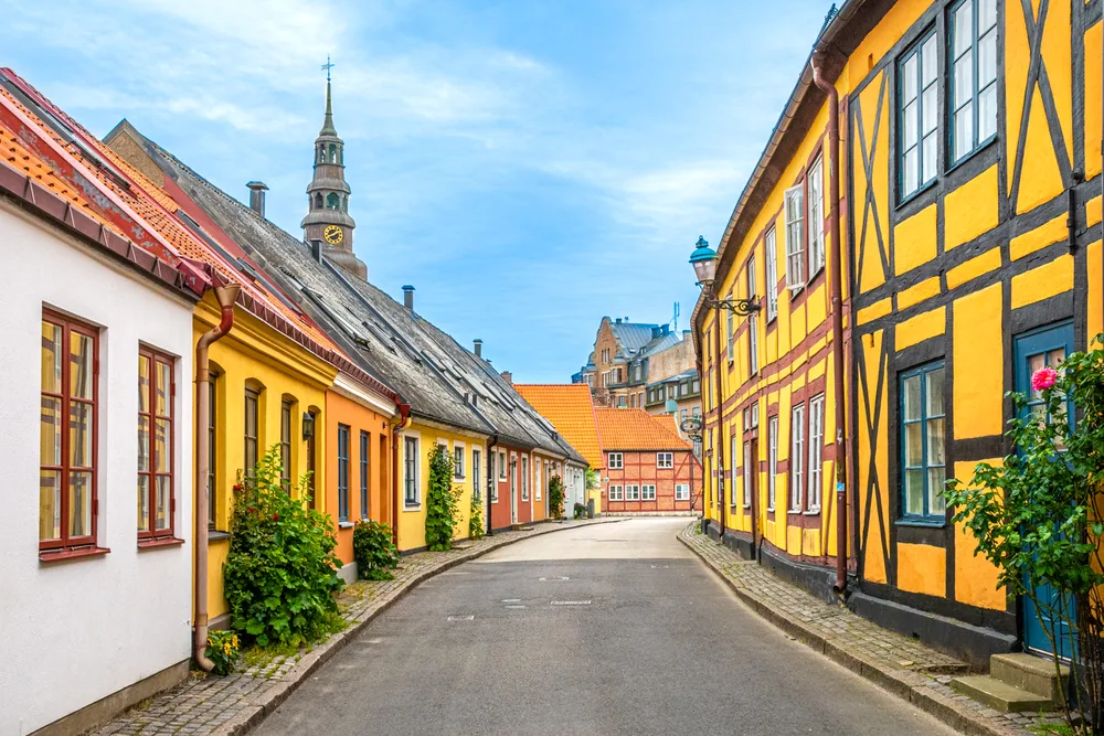 Beautiful day in Ystad Skane Sweden pictured for a guide titled Is Sweden Safe to Visit, with its yellow and brown trimmed buildings on either side of a narrow cement road with a church's steeple towering above the scene