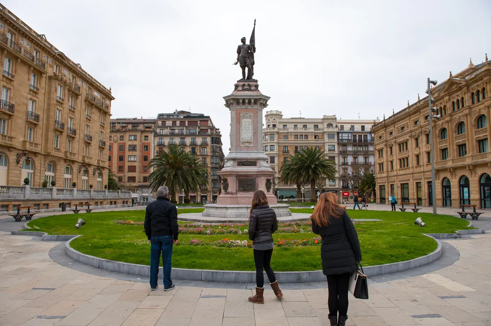 Three people standing in front of a statue situated in the middle of historical structures. 