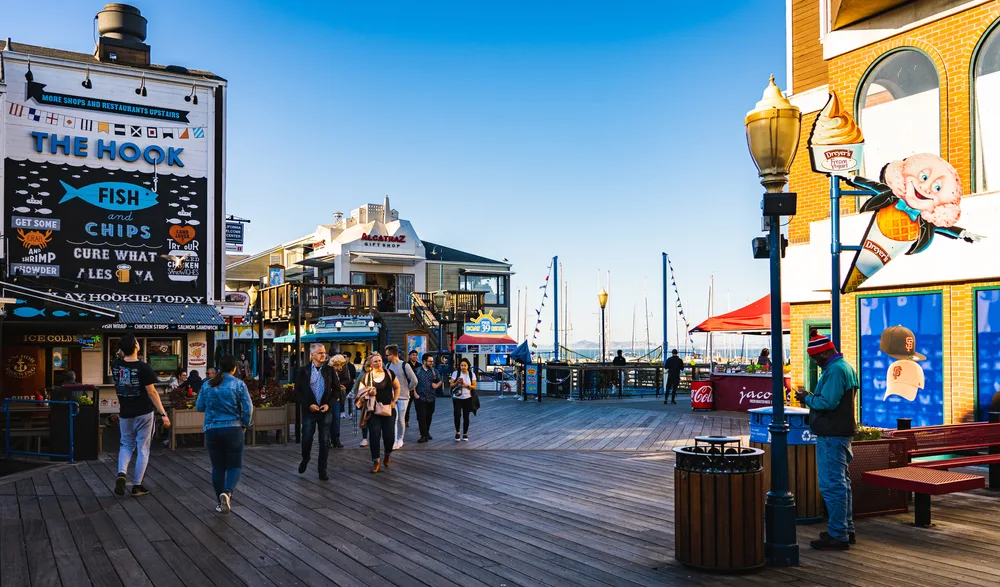Tourists walking on a wood board-covered area, beside a ship port, where various restaurants can be seen in background. 