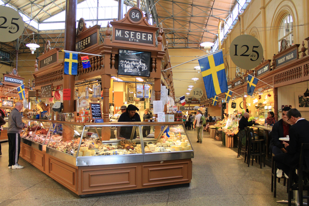 A pastry store with hanging Swedish flag inside the market building, people are sitting on the chair of other stalls in the market, captured for a piece on trip to Sweden cost.