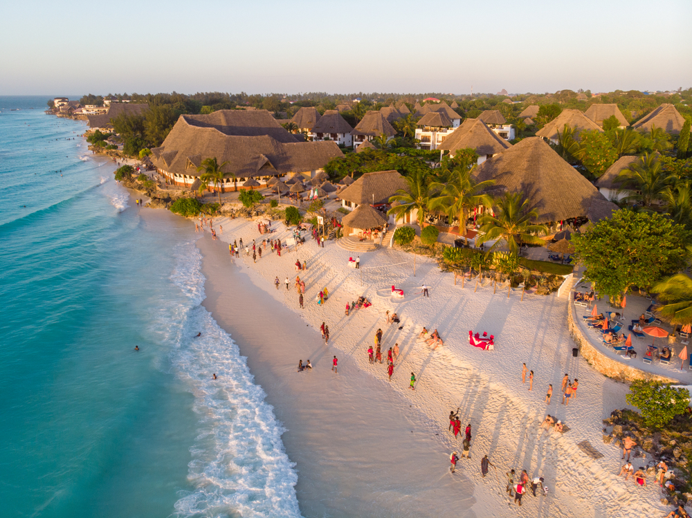 Aerial view of a lovely beach with people enjoying the shore of Nungwi during sunset, one of the best places to stay in Zanzibar; hotels with native roofs can be seen offshore. 