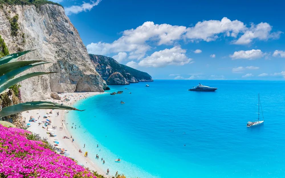 Landscape of Porto Katsiki Beach pictured for a guide titled Is Greece Safe to Visit, with a giant towering cliff on the left and a teal and dark blue beach on the right