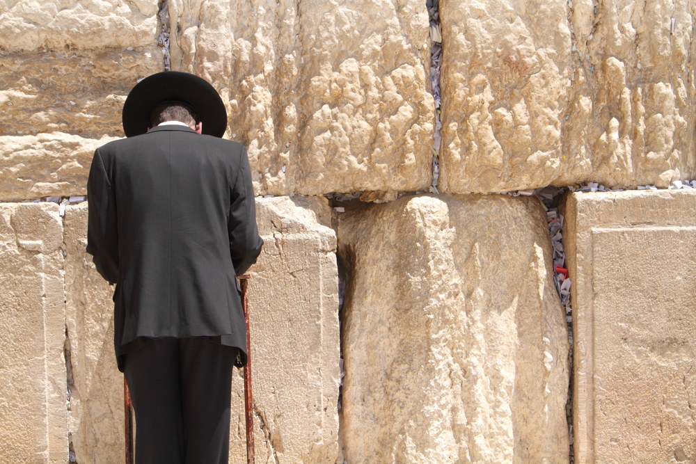 A man wearing a boat and a hat appears to be praying while faching a wall made of huge rocks. 