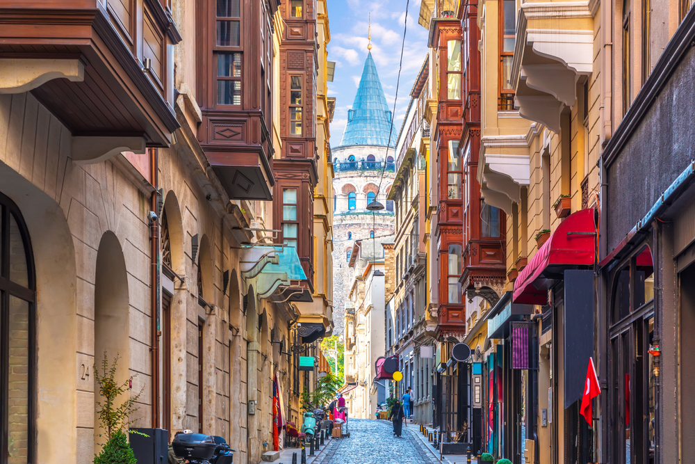 Picturesque view of the Galata Tower in Istanbul pictured from a narrow brick street and framed by tall and historic buildings that house little shops, with a few conservatively-dressed woman walking down the street