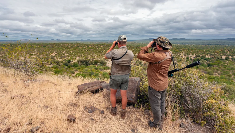 Two men wearing hats while using binoculars and one of them is seen with a long hunting rifle strapped on his shoulder. 