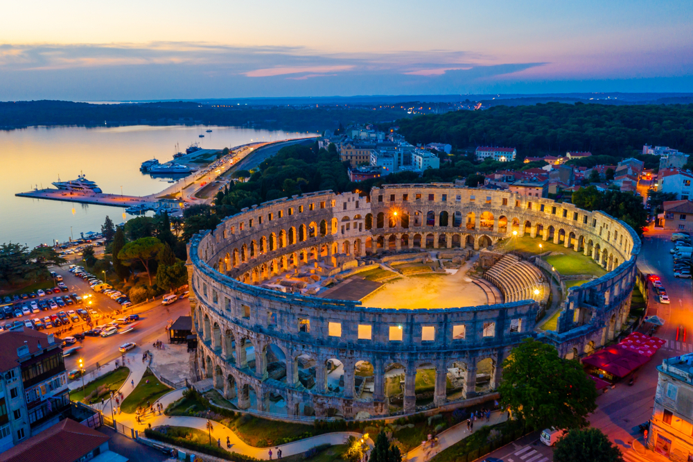 A historical amphitheatre preserved through time in Pula, one of the best areas to stay in Croatia, seats in a city surrounded by modern structures, and a calm bay in can be seen in background during sunset. 