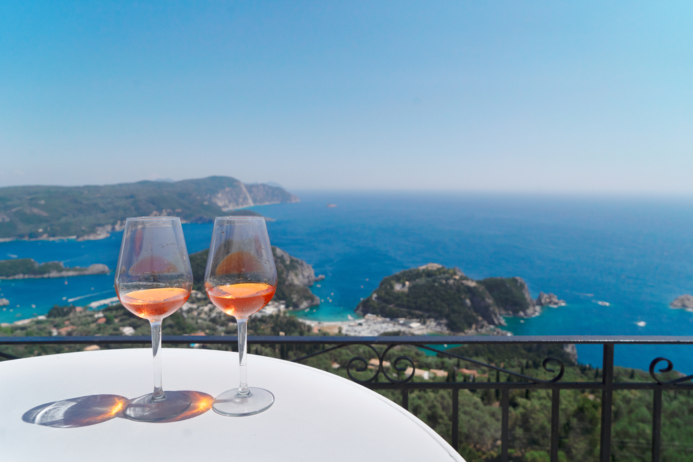 Two wineglass filled with wine is put on a white table, and in background is a beautiful view of the beaches. 