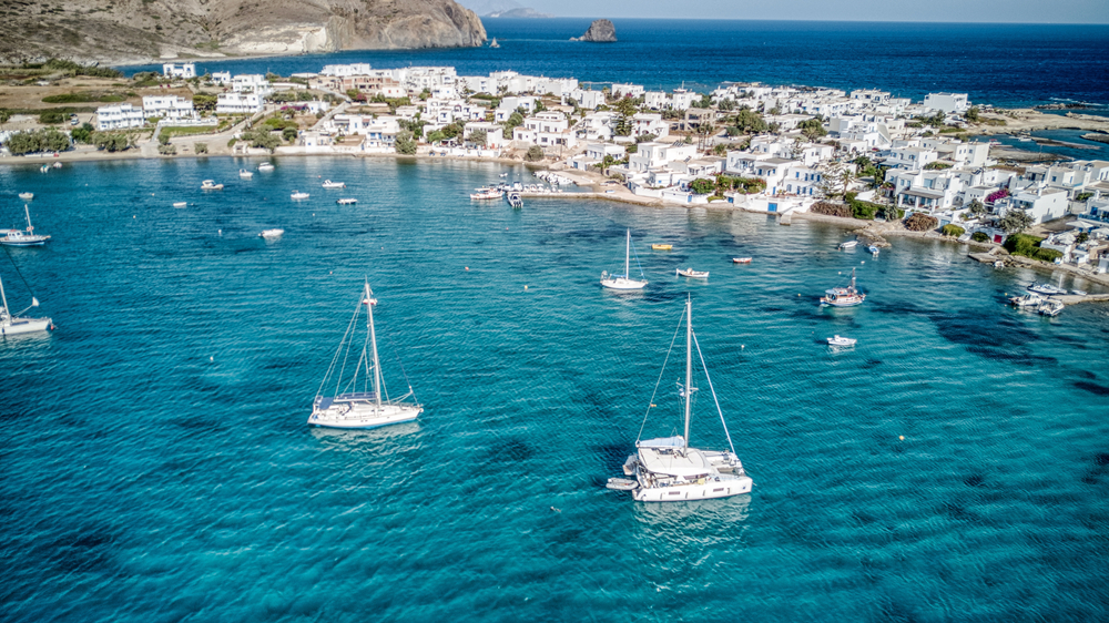 Sailboats moored at a distance to a small village with white structures, an image for a travel guide about safety in visiting Milos.