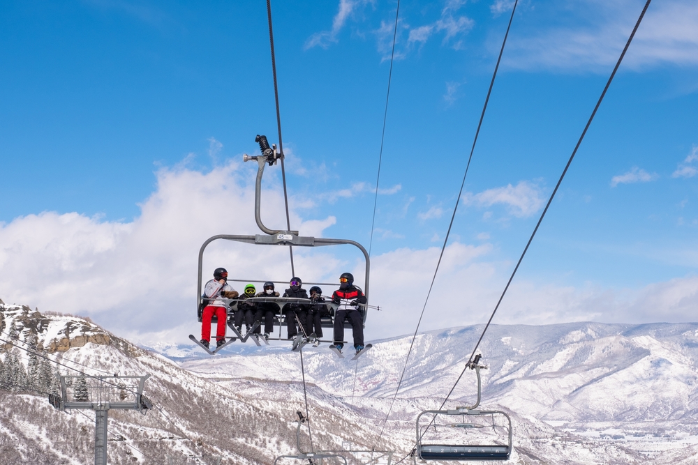A family excitedly rides a ski chairlift going up, and a snowy mountain in background.