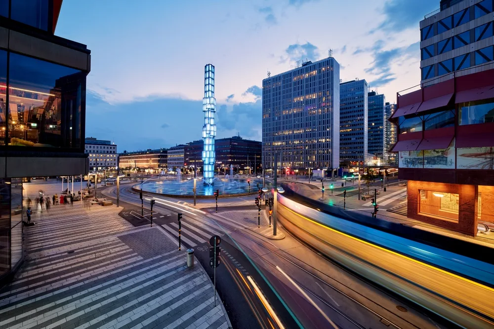 Photo of blue and yellow trams making their way by the photographer in a long exposure image showing a great deal of traffic making its way past the Sergels square intersection with its tall monument in the middle