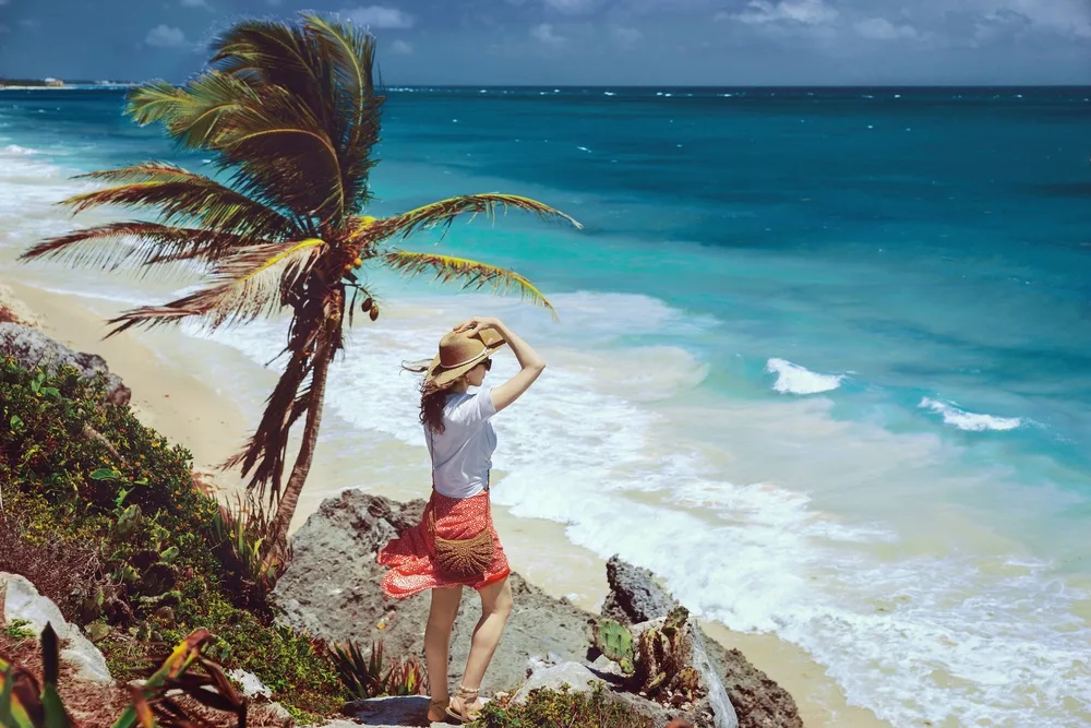 A woman holding her summer hat, standing on a rock at the shore while facing the raging waves on the beach. 