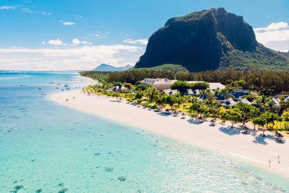 A tropical destination with white sand beach in Le Morne, one of the best areas to stay in Mauritius, a tall land formation covered with lush forest is at the middle of the area, while hotel structures stand behind the palm trees on the shore.