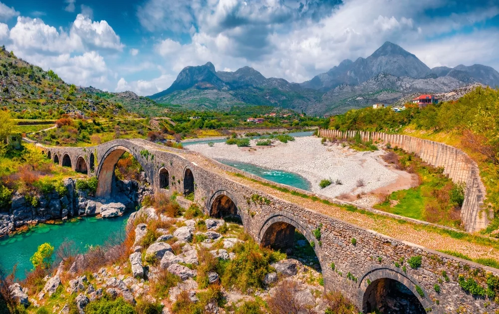 Old Mes Bridge in Albania seen with the river running under it and cloudy blue skies overhead for a list of the cheapest places to travel in Europe