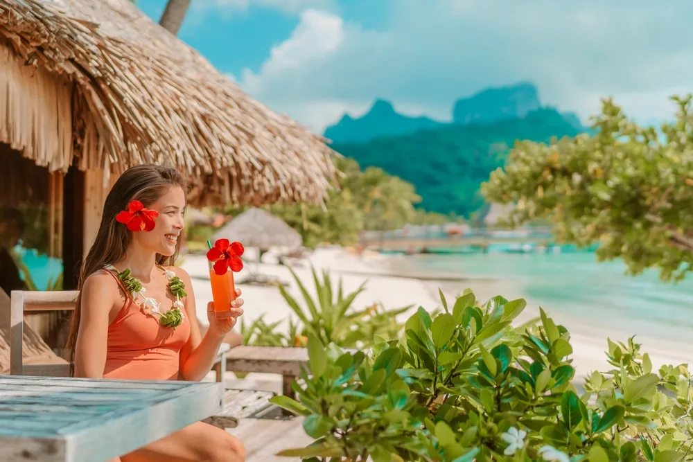A woman sitting on a chair beside a native hut by the beach while smiling and holding a tropical drink, an image for a travel guide about trip to Tahiti cost.
