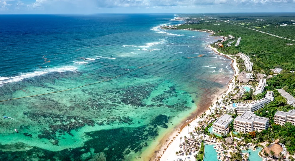 Aerial view on a long beach where hotels can be seen beside crystal clear waters, white sand, and trees off shore. 