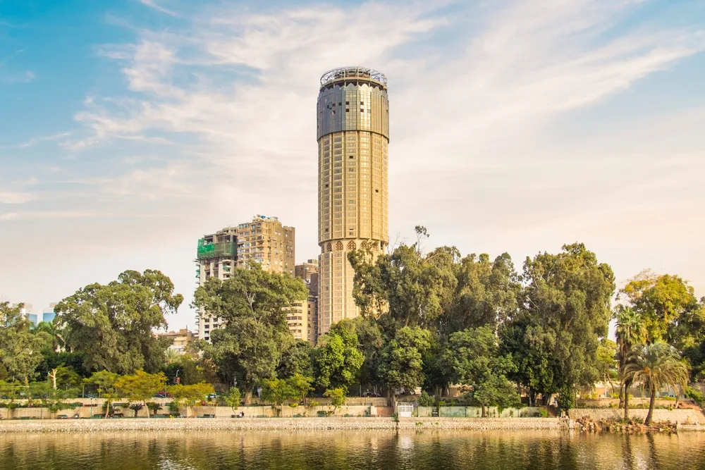 A view from the river in Zamalek, one of the best areas to stay in Cairo, the other of the riverbank are filled with lush trees and at distant is a tall cylindrical building in the city proper.