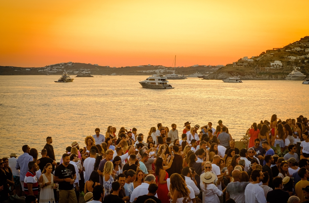 People partying in the beach-side during sunset, an image for a travel guide about safety in visiting Mykonos, boats can be see cruising in the see. 