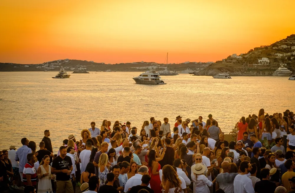People partying in the beach-side during sunset, an image for a travel guide about safety in visiting Mykonos, boats can be see cruising in the see. 