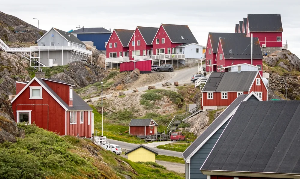 To illustrate that Greenland is very safe to visit, a bunch of red and blue homes sit on a cliff in a small village with rocky and grassy hills