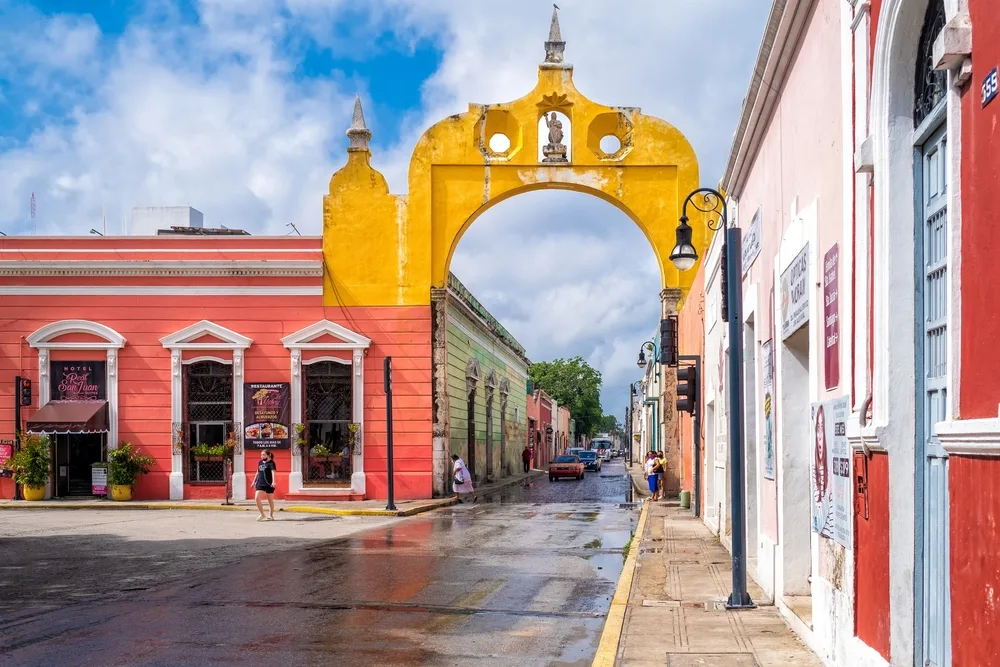 An old golden arc structure sits above the wet street of Centro Historico, and people pass by below it.